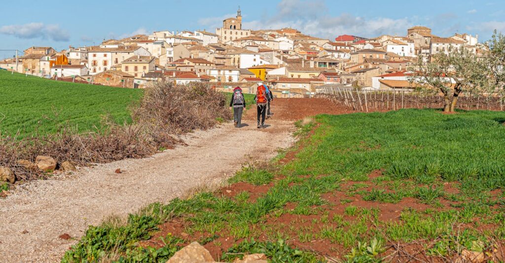 Two pilgrims with backpacks walk towards the picturesque village of Cirauqui, Spain, on a sunny day.